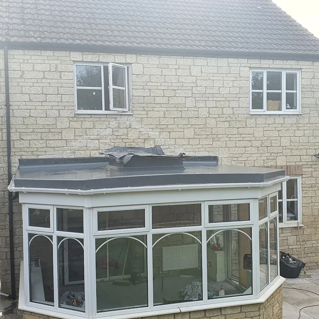 Bay-shaped conservatory with a new solid grey flat roof fitted on top of the frame and a rooflight still wrapped in protective sheeting